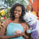 Michelle Obama, wearing a blue apron, stands in a rooftop garden posing for the camera and holding a pink, round mochi puppet. A bee puppet wearing a red tie hovers over her shoulder while a furry white and blue puppet with frozen waffle ears embraces her side.