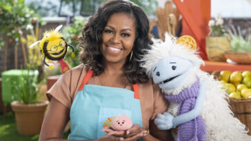Michelle Obama, wearing a blue apron, stands in a rooftop garden posing for the camera and holding a pink, round mochi puppet. A bee puppet wearing a red tie hovers over her shoulder while a furry white and blue puppet with frozen waffle ears embraces her side.